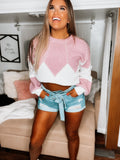 A woman wearing a pink color block sweater with pearl beading, paired with blue denim shorts.