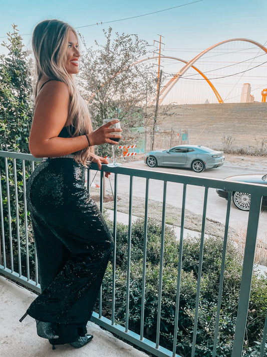 A person standing on a balcony wearing loose sparkle sequin black trousers with an elasticated waistband, paired with a black top and drinking from a glass.