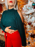A person standing in front of a Christmas tree wearing a black turtleneck, shoulders open sweater and red pants.