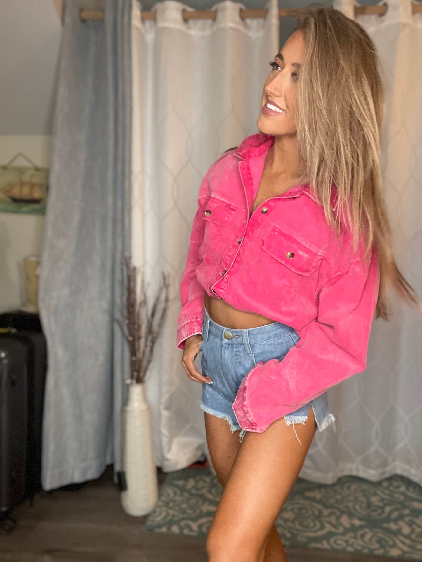 A person wearing a pink denim jacket with a round neckline and long sleeves, paired with blue denim shorts. The setting appears to be indoors, possibly in a home environment.
