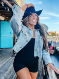 A woman wearing a black sport romper and a cowboy hat, standing on a deck with a body of water and buildings in the background.
