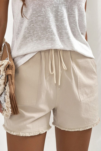 A pair of light beige, drawstring shorts are shown. 

The shorts are made of a slightly textured fabric. They have a drawstring waist with light beige/cream colored drawstrings. 

There are small patch pockets on each side. The hemline has a frayed, slightly uneven edge.

A light, off-white, slightly sheer, loose-fitting t-shirt or top is visible underneath the shorts. A woven bag or purse with a fringe detail in beige and brown is partially visible in the lower left of the image.