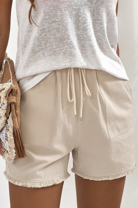 A pair of light beige, drawstring shorts are shown. 

The shorts are made of a slightly textured fabric. They have a drawstring waist with light beige/cream colored drawstrings. 

There are small patch pockets on each side. The hemline has a frayed, slightly uneven edge.

A light, off-white, slightly sheer, loose-fitting t-shirt or top is visible underneath the shorts. A woven bag or purse with a fringe detail in beige and brown is partially visible in the lower left of the image.
