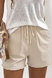 A pair of light beige, drawstring shorts are shown. 

The shorts are made of a slightly textured fabric. They have a drawstring waist with light beige/cream colored drawstrings. 

There are small patch pockets on each side. The hemline has a frayed, slightly uneven edge.

A light, off-white, slightly sheer, loose-fitting t-shirt or top is visible underneath the shorts. A woven bag or purse with a fringe detail in beige and brown is partially visible in the lower left of the image.
