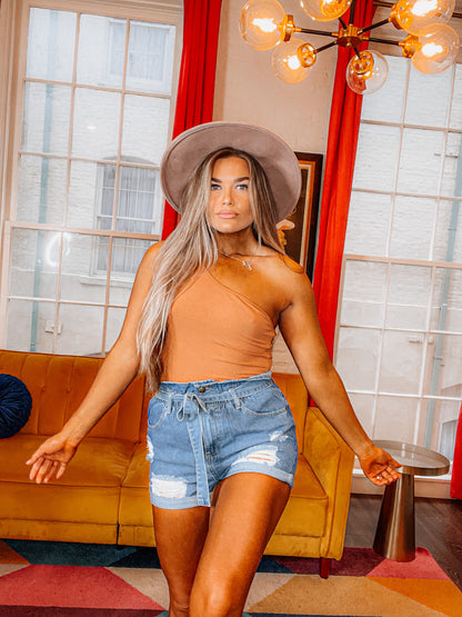 A person wearing high-waisted blue denim shorts with ripped detailing, paired with an orange top and a brown hat.
