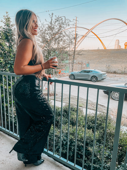 A person standing on a balcony wearing loose sparkle sequin black trousers with an elasticated waistband, paired with a black top and drinking from a glass.