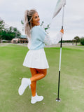 A woman in a golf course holding a golf club and wearing a white skirt with a sweater and white shoes. The background is green, indicating the grassy area of a golf course.