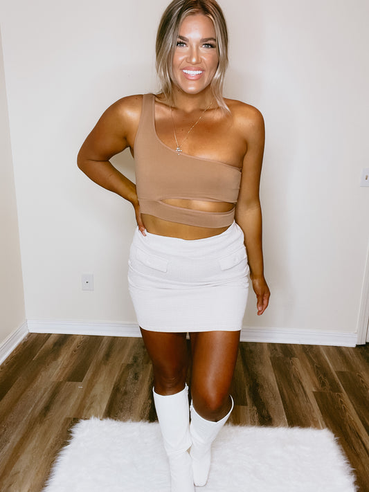 A person wearing a brown sleeveless top and a beige skirt, standing in a room with wooden flooring.