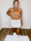 A person wearing a brown sleeveless top and a beige skirt, standing in a room with wooden flooring.