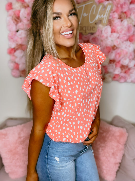 A person wearing a vibrant peach hue top with short sleeves and a round neckline, standing in a room with pink decor.